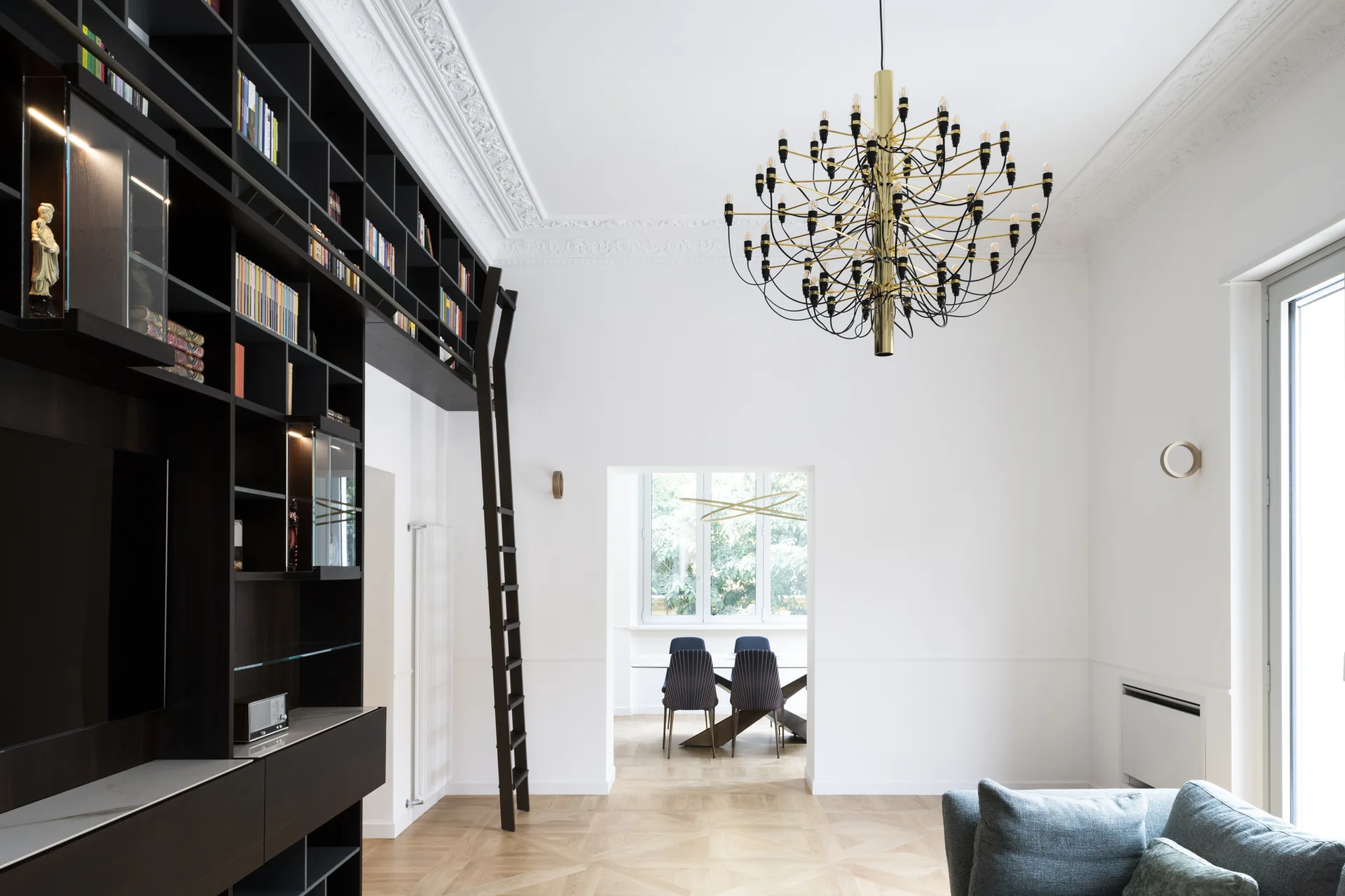 A bright white living room features a dark built-in bookshelf with a ladder, a brass multi-arm chandelier, and parquet floors.