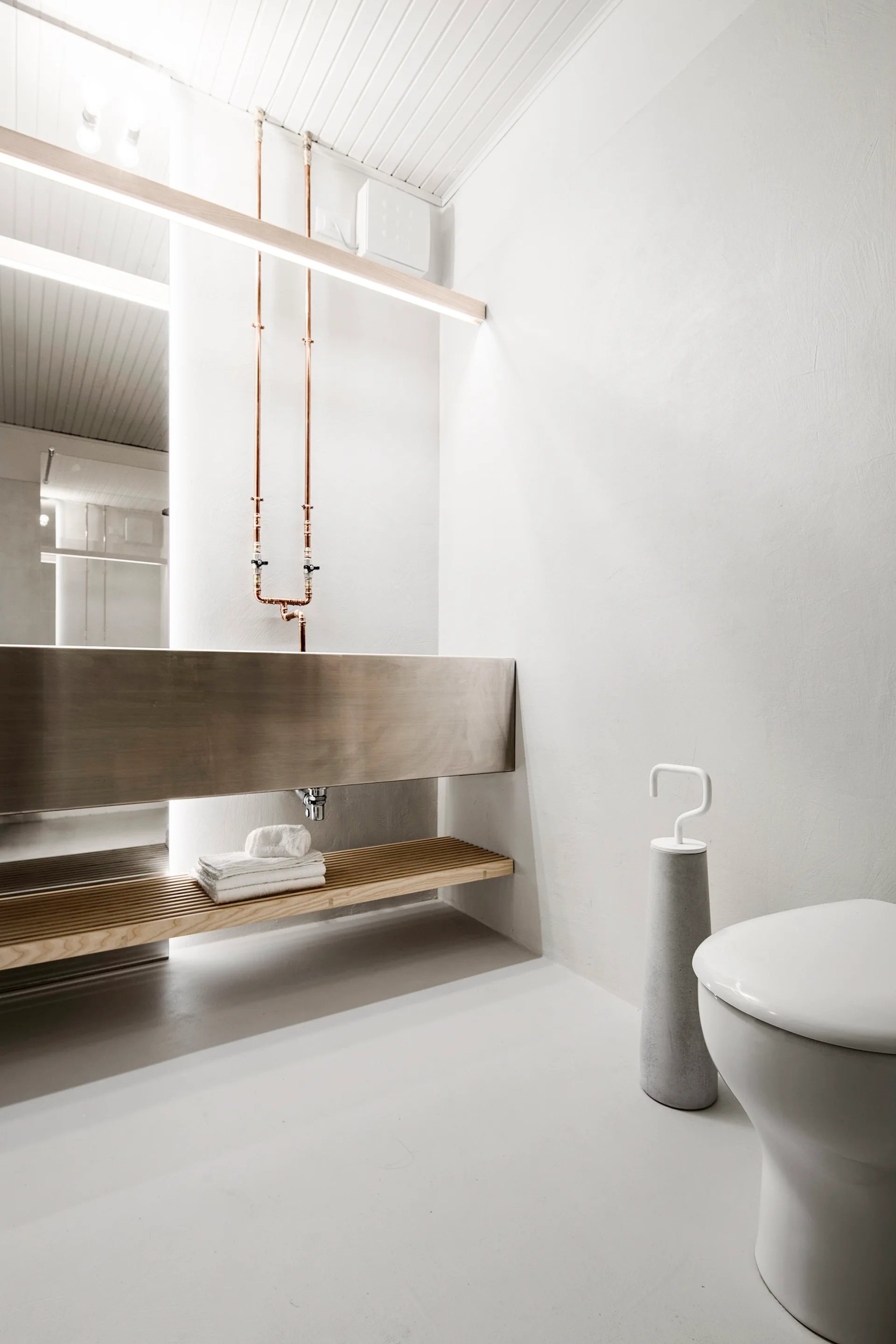 A minimalist white bathroom featuring a wall-mounted metal trough sink, exposed copper pipes, a slatted wooden shelf, and a white toilet.