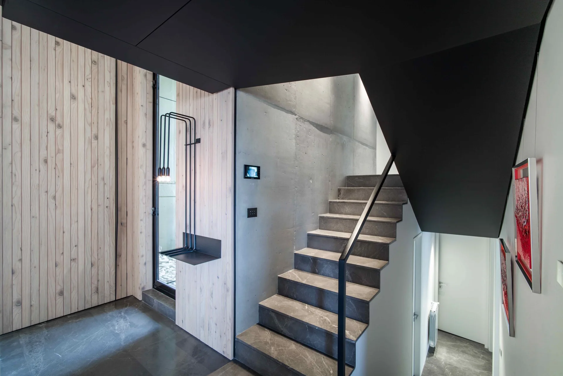A gray stone staircase with a slim black handrail set between a raw concrete wall and vertical wood paneling.