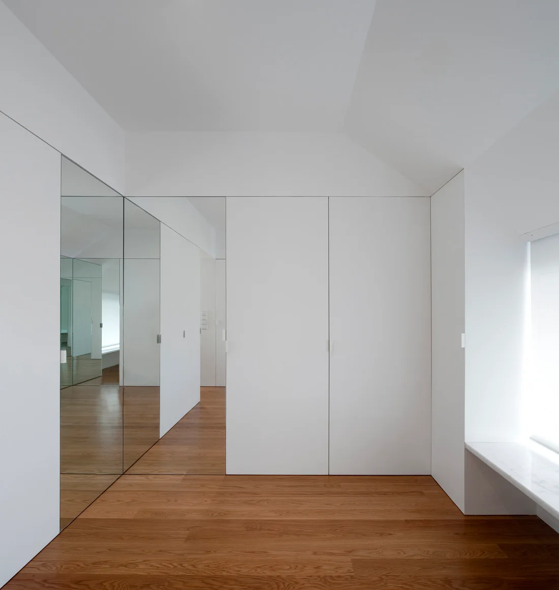 A minimalist room with built-in white wardrobes, a full-height mirrored wall panel, and warm wood flooring under a sloped ceiling.