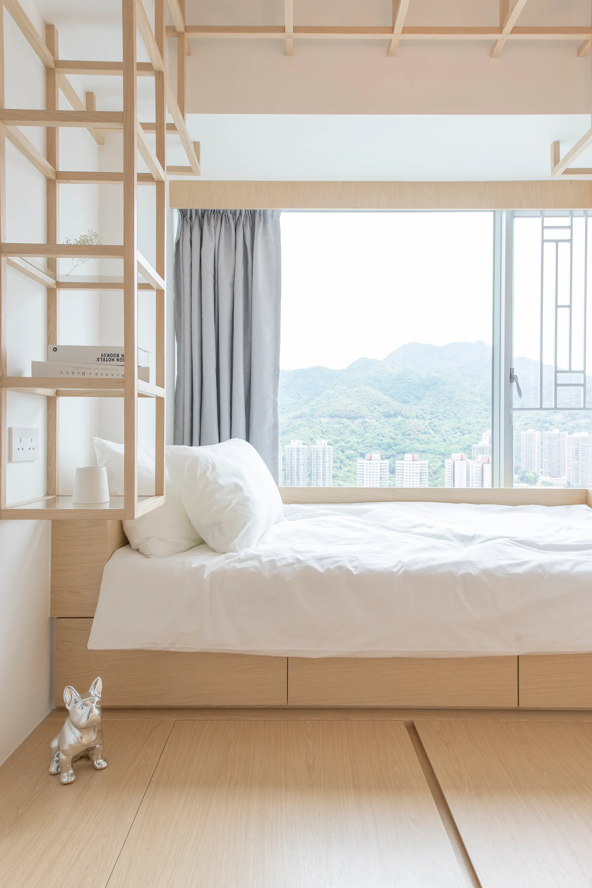A minimalist bedroom featuring a light wood platform bed with white bedding, an open ladder shelving unit, and a decorative wood ceiling grid.