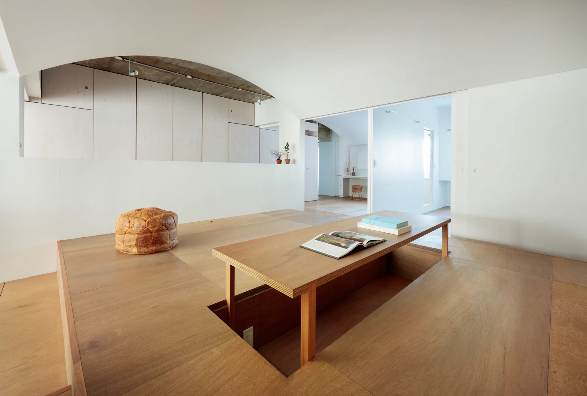 A minimalist room features a plywood platform floor with a sunken seating well and table, a brown leather pouf, and a curved white ceiling.