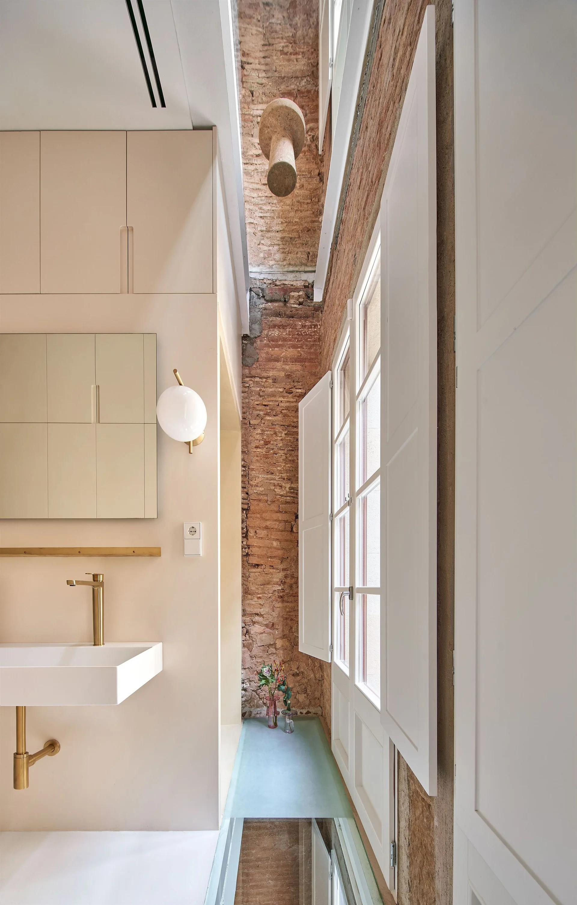 A minimalist bathroom vanity area with a pale pink sink cabinet, modern gold faucet, and globe sconce set against an exposed brick wall.