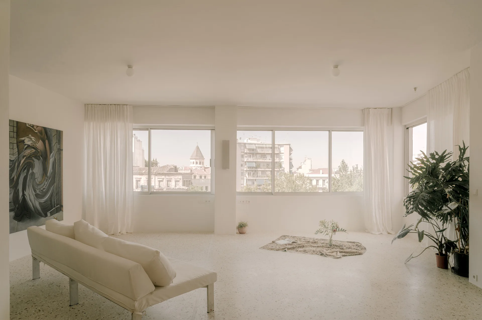 A minimalist white living room features a low-profile white sofa, speckled terrazzo flooring, and large windows with sheer drapes.