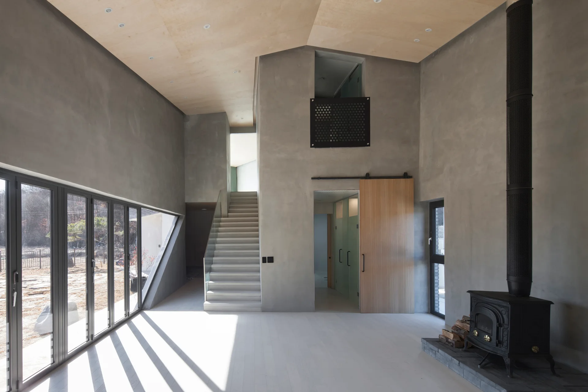 A minimalist double-height living room featuring gray concrete walls, a sloped plywood ceiling, a concrete staircase, and a black wood-burning stove.