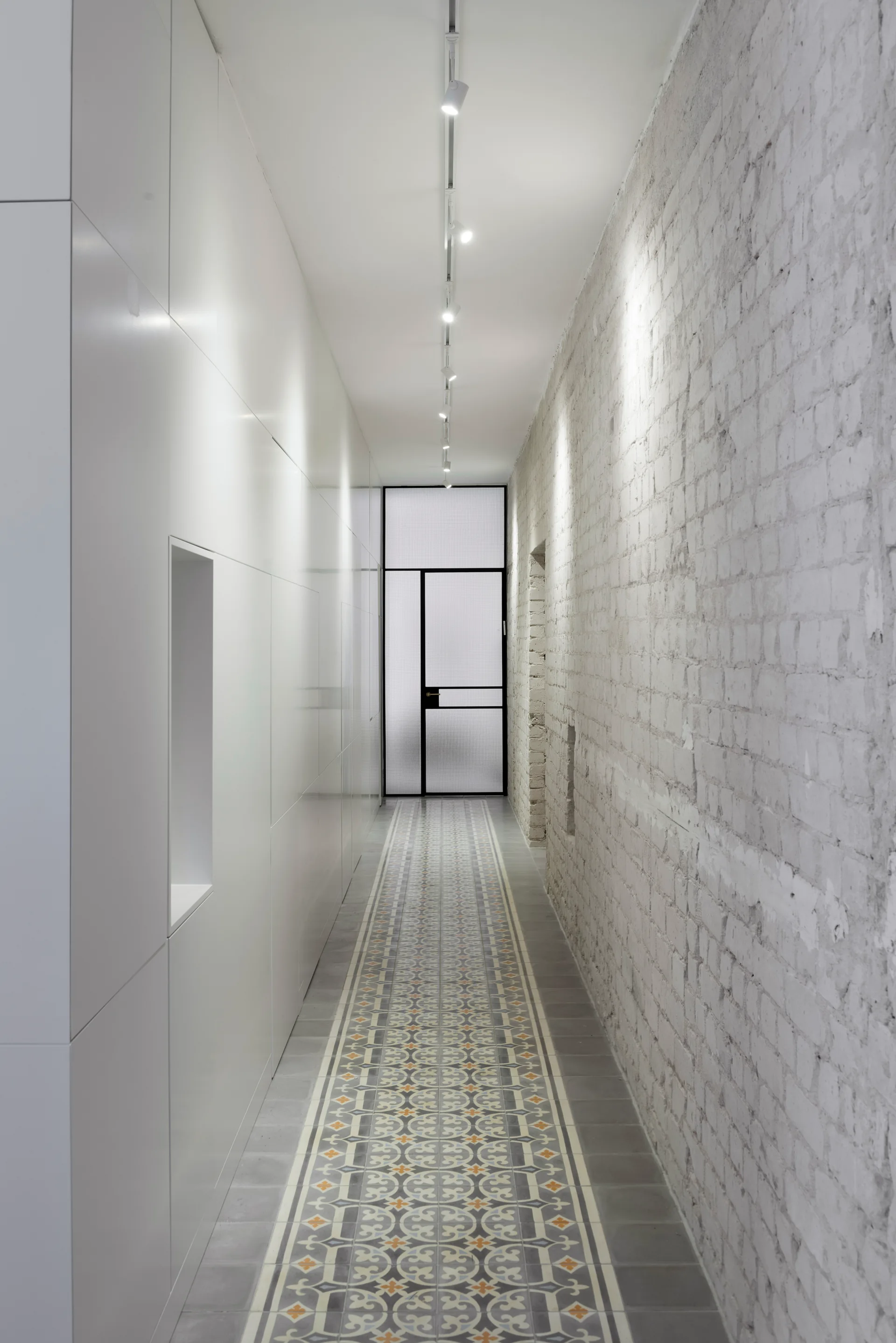 A narrow hallway features a patterned tile floor runner between a smooth white paneled wall and a white painted brick wall.