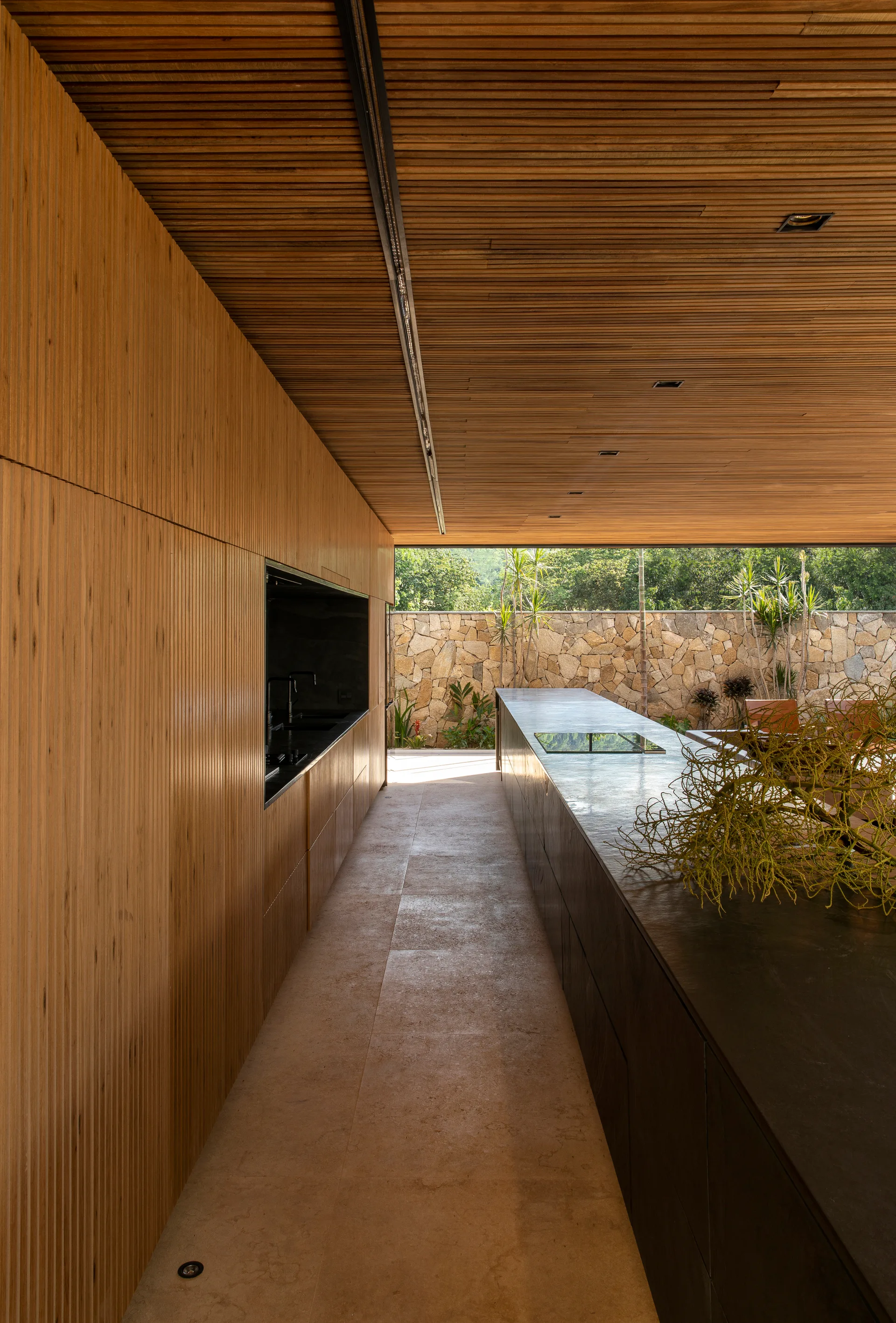 A minimalist kitchen features a long dark stone-like island, a slatted wood ceiling and wall, and a view of an outdoor stone masonry wall.