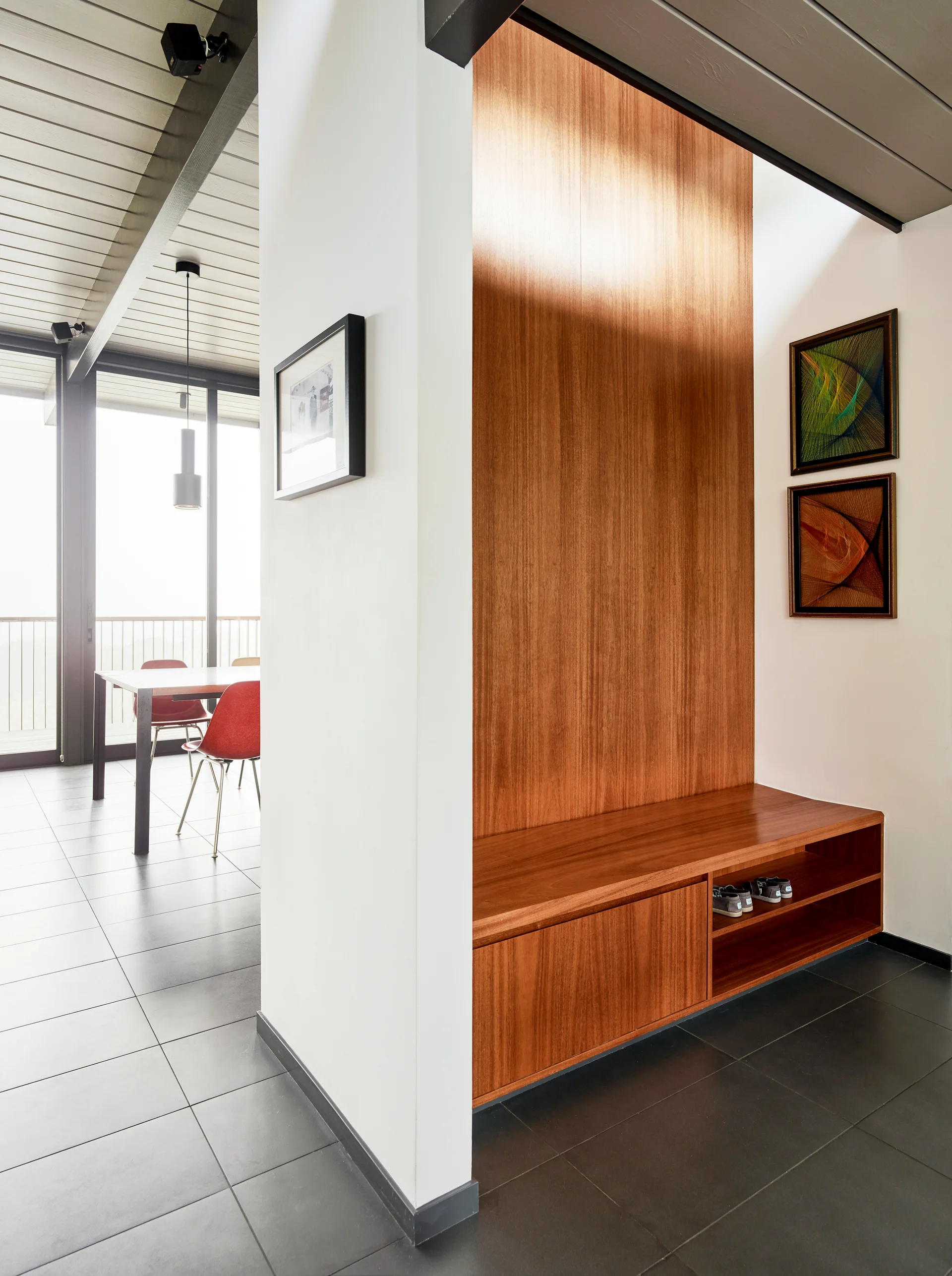 A modern entryway features a warm wood-paneled wall, built-in storage bench, dark tile flooring, and a partition wall looking into a dining area.