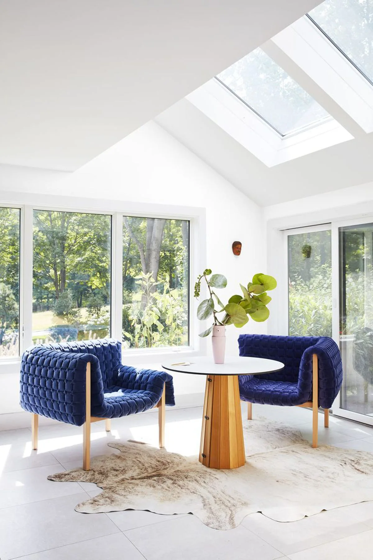 Two blue quilted armchairs flank a wood pedestal table on a cowhide rug in a sunlit room with a skylight.