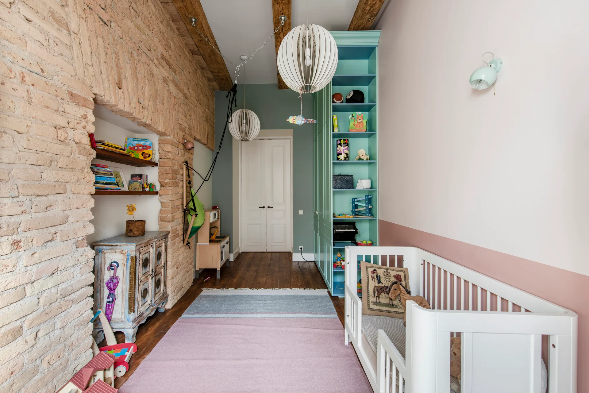 A children's room featuring a white wooden crib, an exposed brick wall, a teal built-in cabinet, and soft pink and green painted walls.