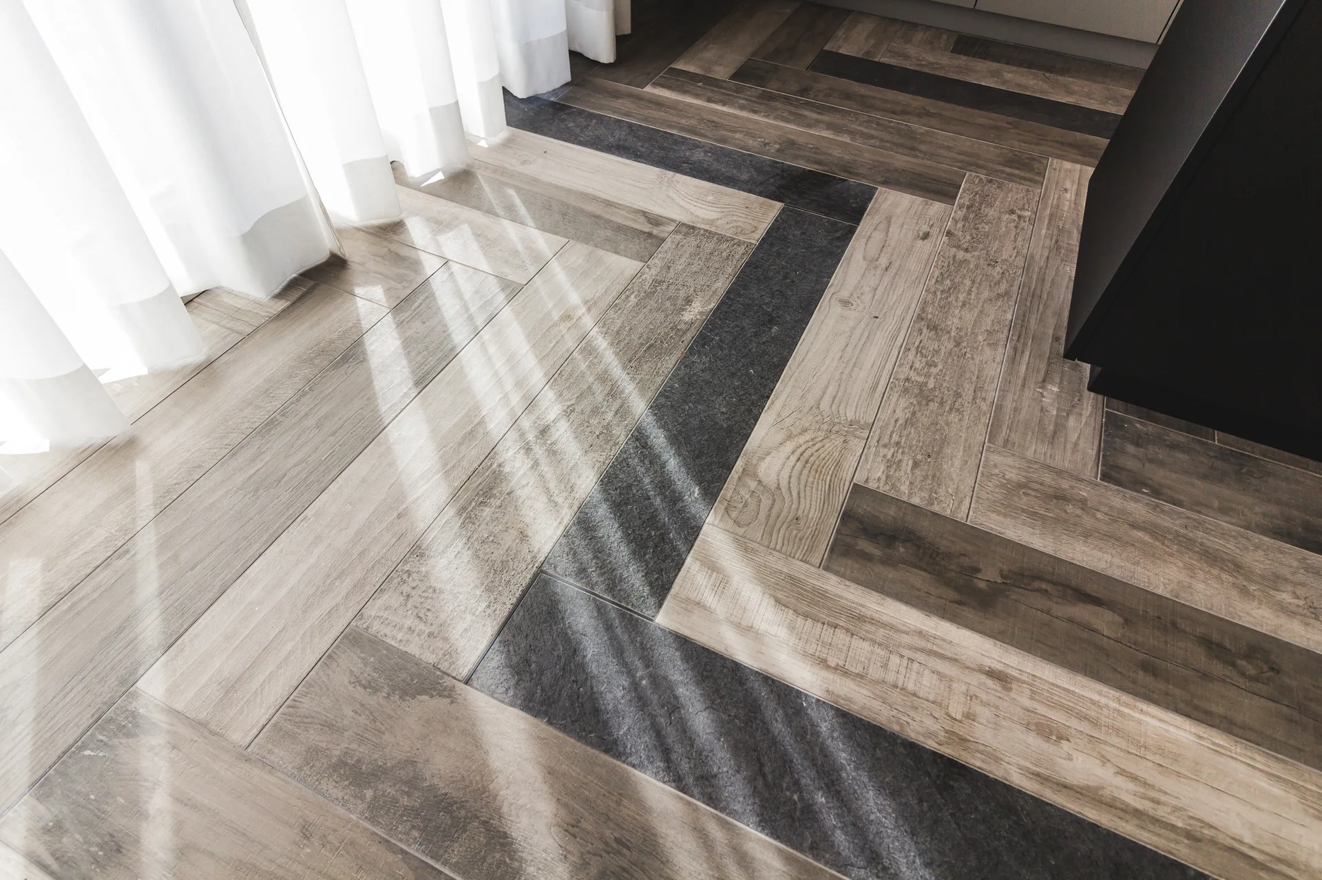 Herringbone patterned wood-look floor tiles with a dark grey accent stripe, illuminated by natural light filtering through sheer white curtains.