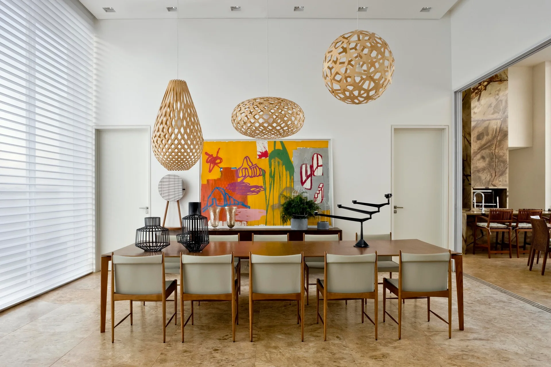 A long wooden dining table with six chairs sits below three woven lattice pendant lights, with a colorful abstract painting on the back wall.