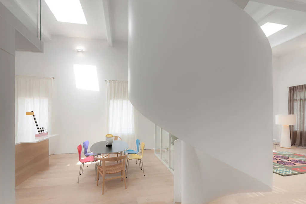 An open concept room features a large white curved sculptural staircase next to an oval dining table with colorful chairs and light wood floors.