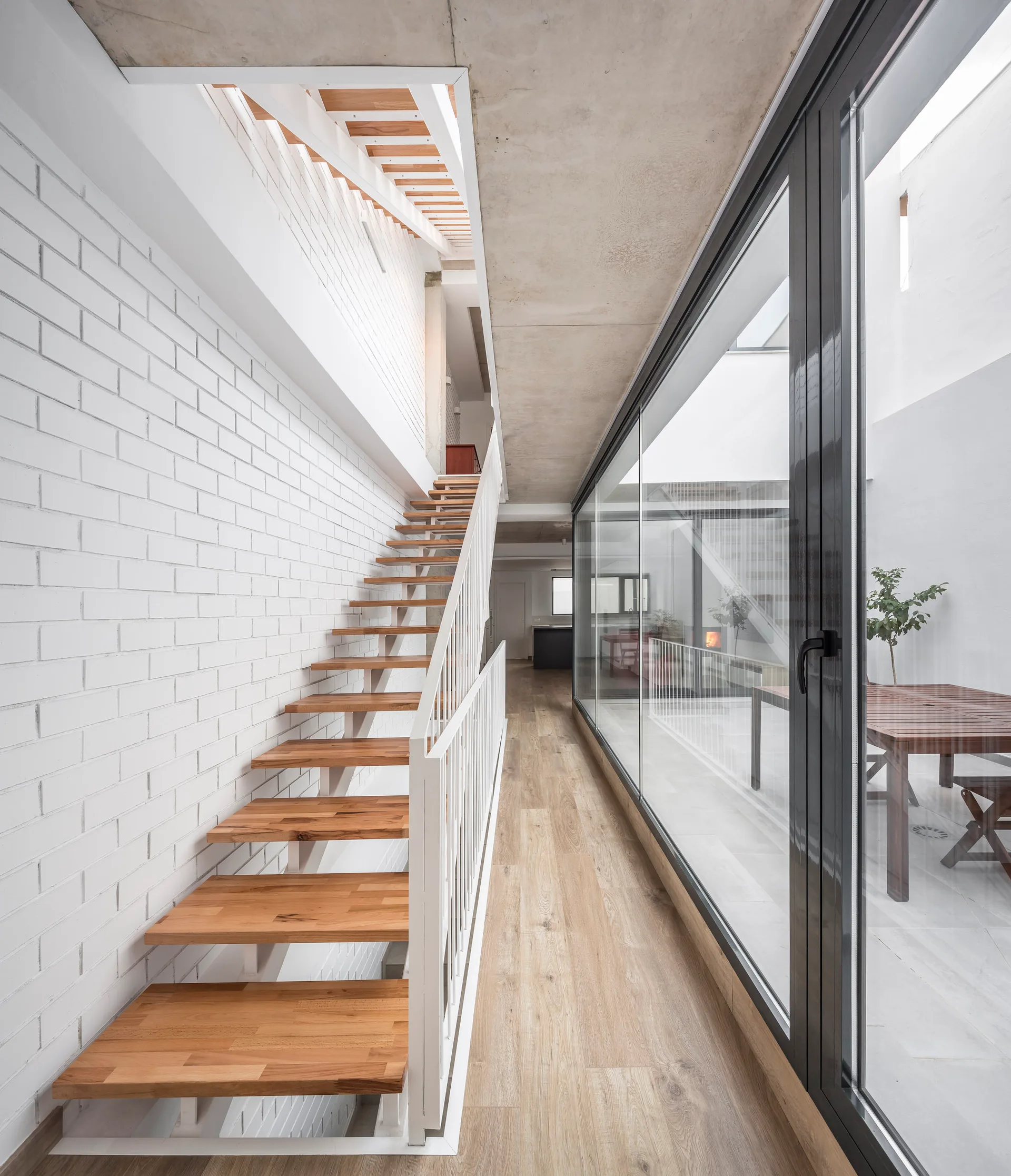A floating wooden staircase with white railings stands beside a glass wall and a white painted brick wall under a concrete ceiling.