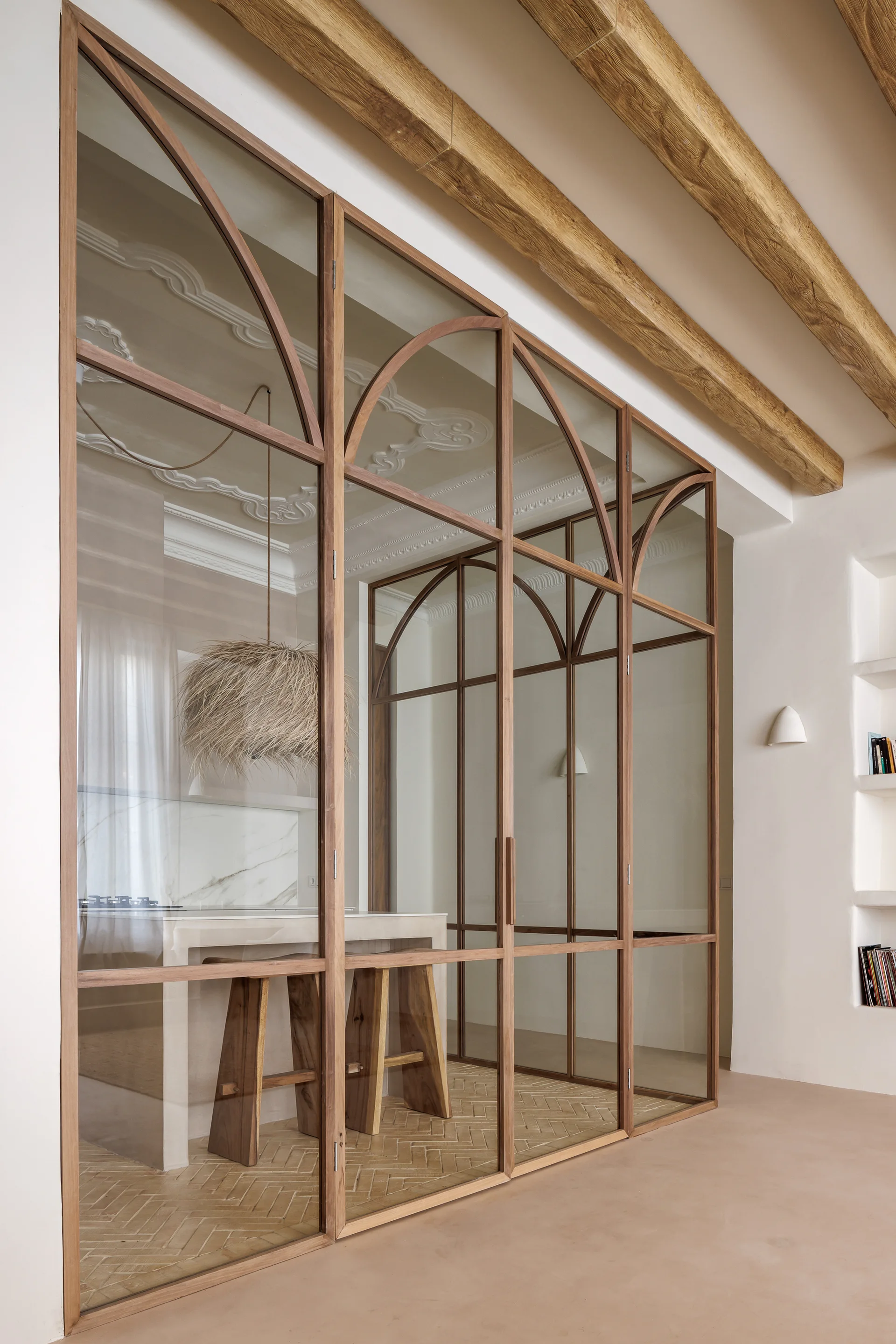 An interior view shows a wood-framed glass partition with arches separating a plaster-walled hallway from a kitchen featuring exposed timber beams and stools.