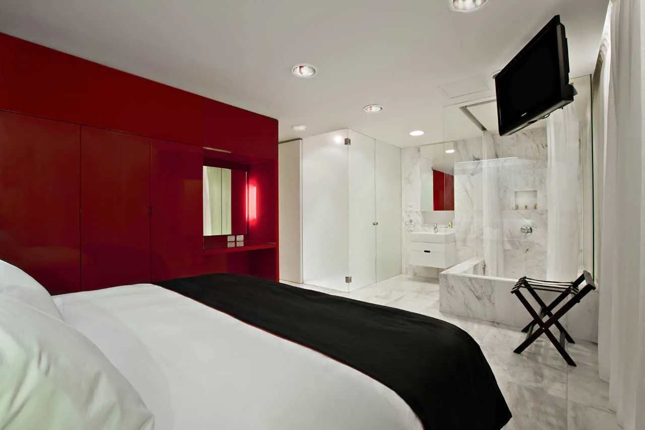 A modern bedroom featuring a bed with white and black bedding, glossy red wardrobes, and an open ensuite bathroom clad in white marble.