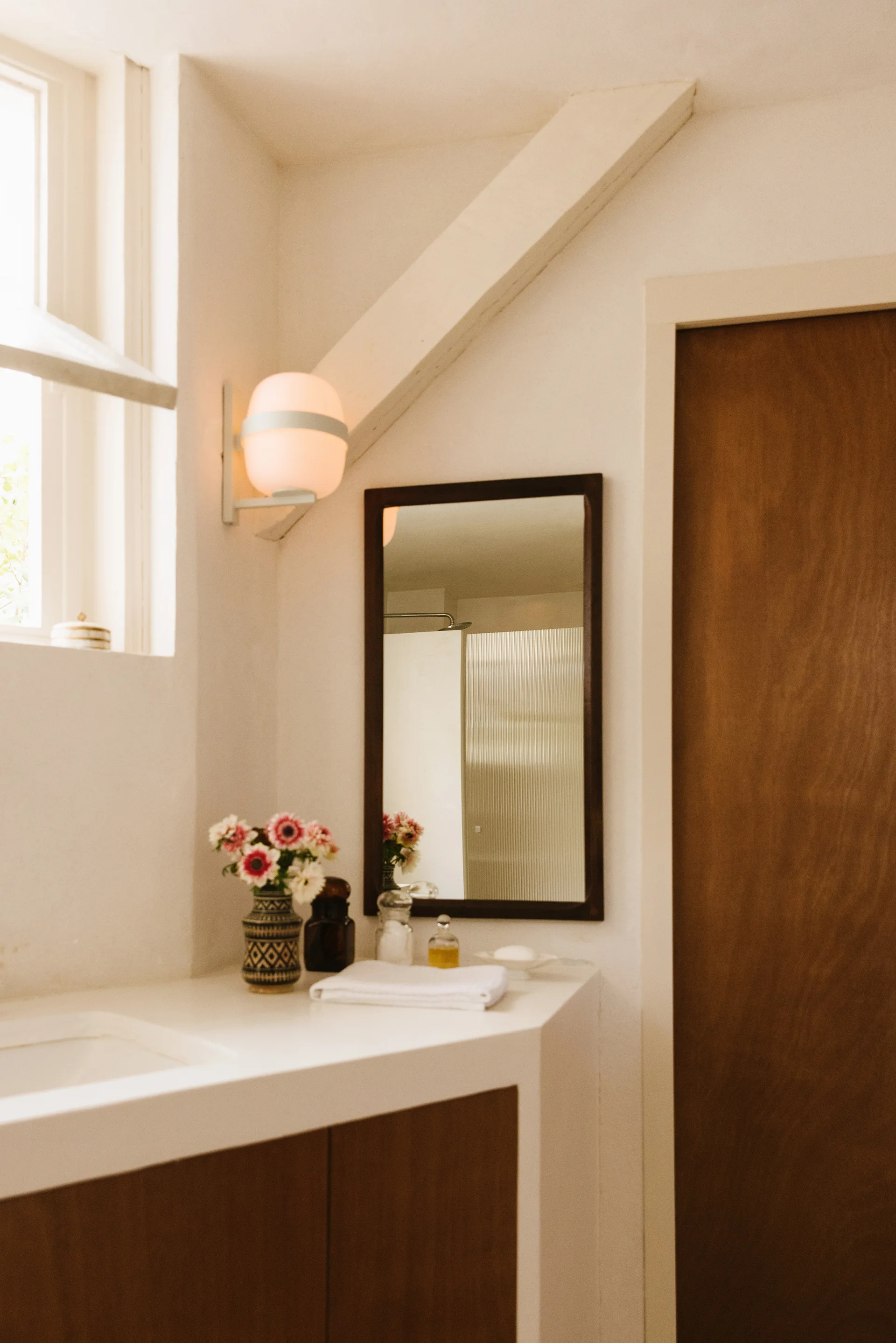 A bathroom vanity with a white countertop and wood cabinet stands beneath a slanted ceiling beam, a rectangular mirror, and a glowing globe sconce.