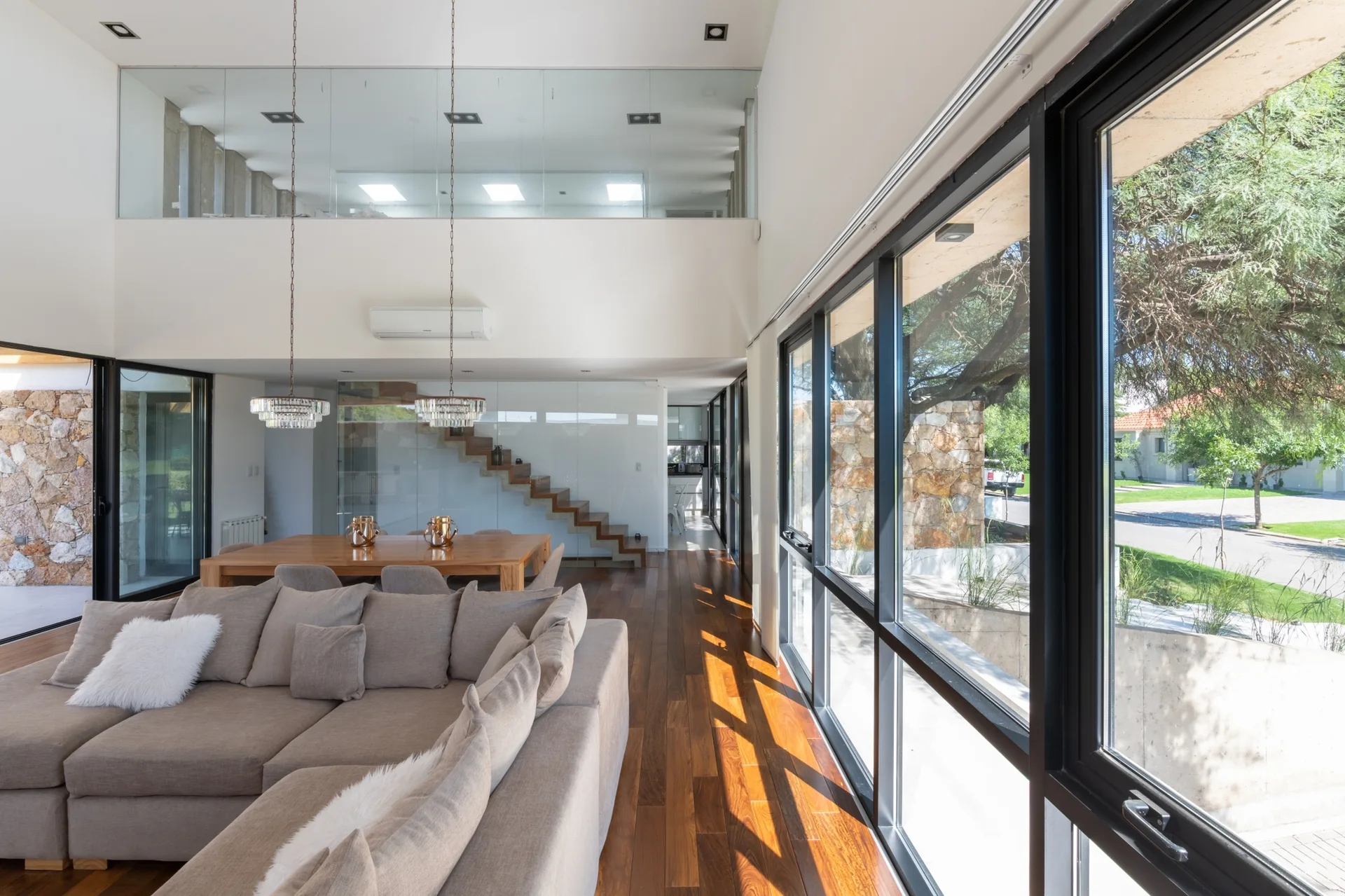 A modern open-plan living room features a tan sectional sofa, polished dark wood floors, and a floating staircase behind a glass partition.