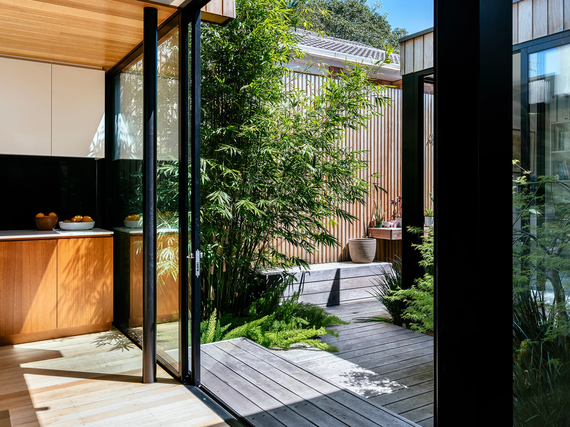 A modern kitchen with wood cabinets opens through sliding glass doors to a sunlit wooden deck with vertical wood slat siding and green bamboo.