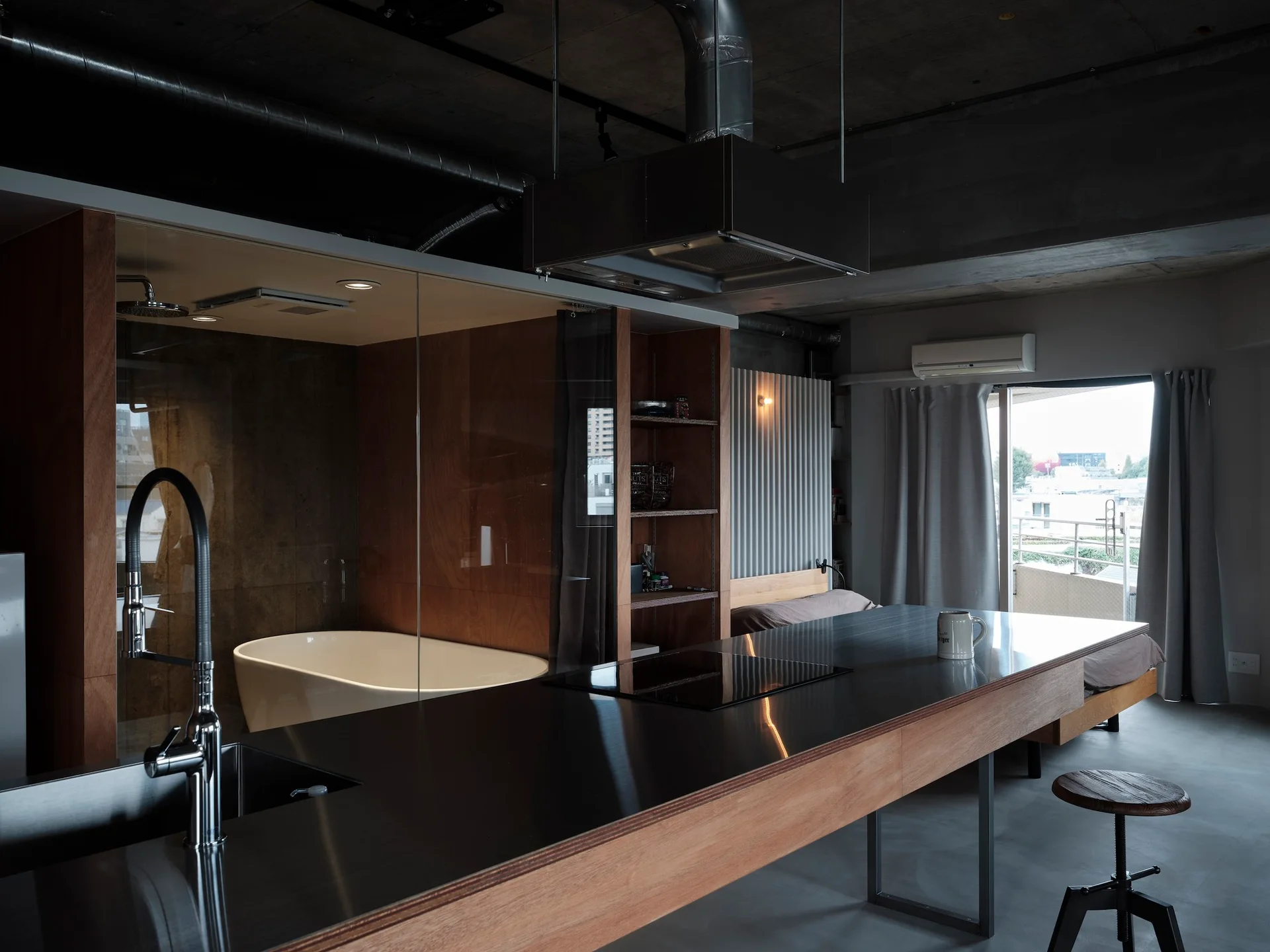 An industrial studio apartment features a long metal kitchen island, a glass-walled bathroom with a bathtub, and an exposed concrete ceiling.