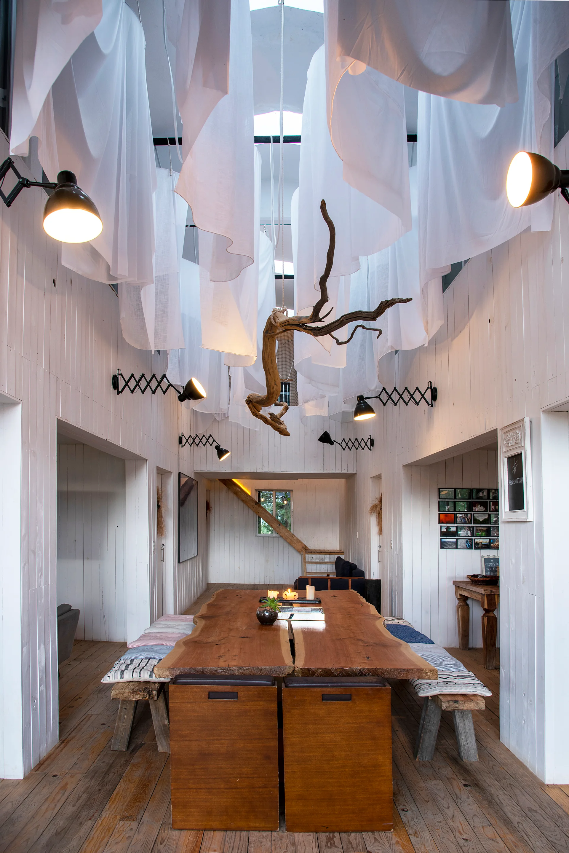 A rustic dining room features a long live-edge wooden table, benches, white plank walls, and white fabric drapery suspended from the ceiling.