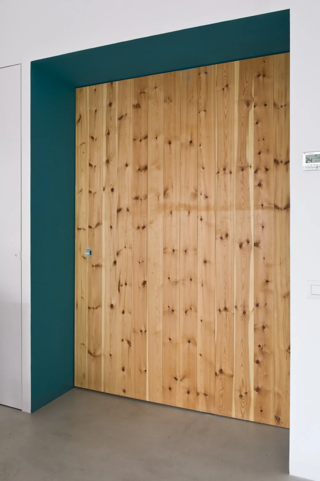 A flush-mounted door made of vertical knotty wood planks is set into a deep teal painted recessed wall niche beside a white wall.