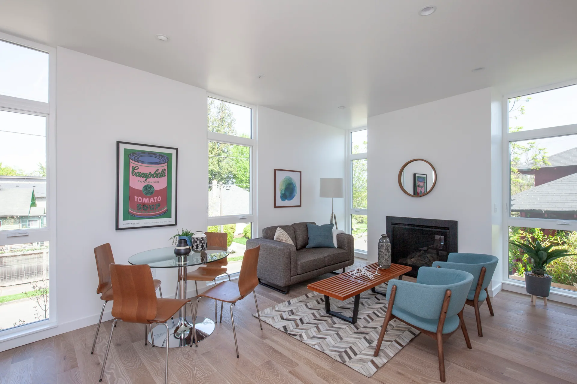 A bright open-concept living space with a glass-top dining table, a grey sofa, teal armchairs, and floor-to-ceiling windows.