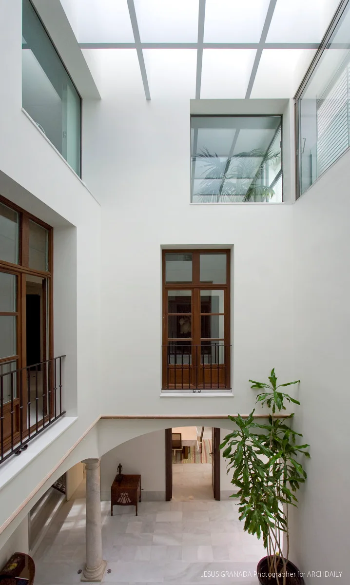 A multi-level white indoor courtyard featuring dark wood-framed windows, a large grid skylight ceiling, stone floors, and a tall green potted plant.