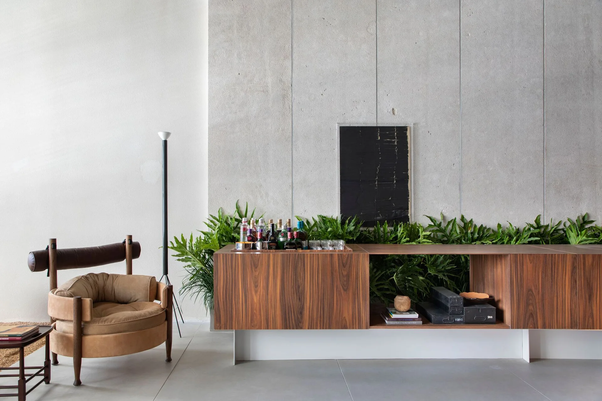 A low wooden bar console with integrated green plants sits against a textured concrete wall beside a tan armchair.