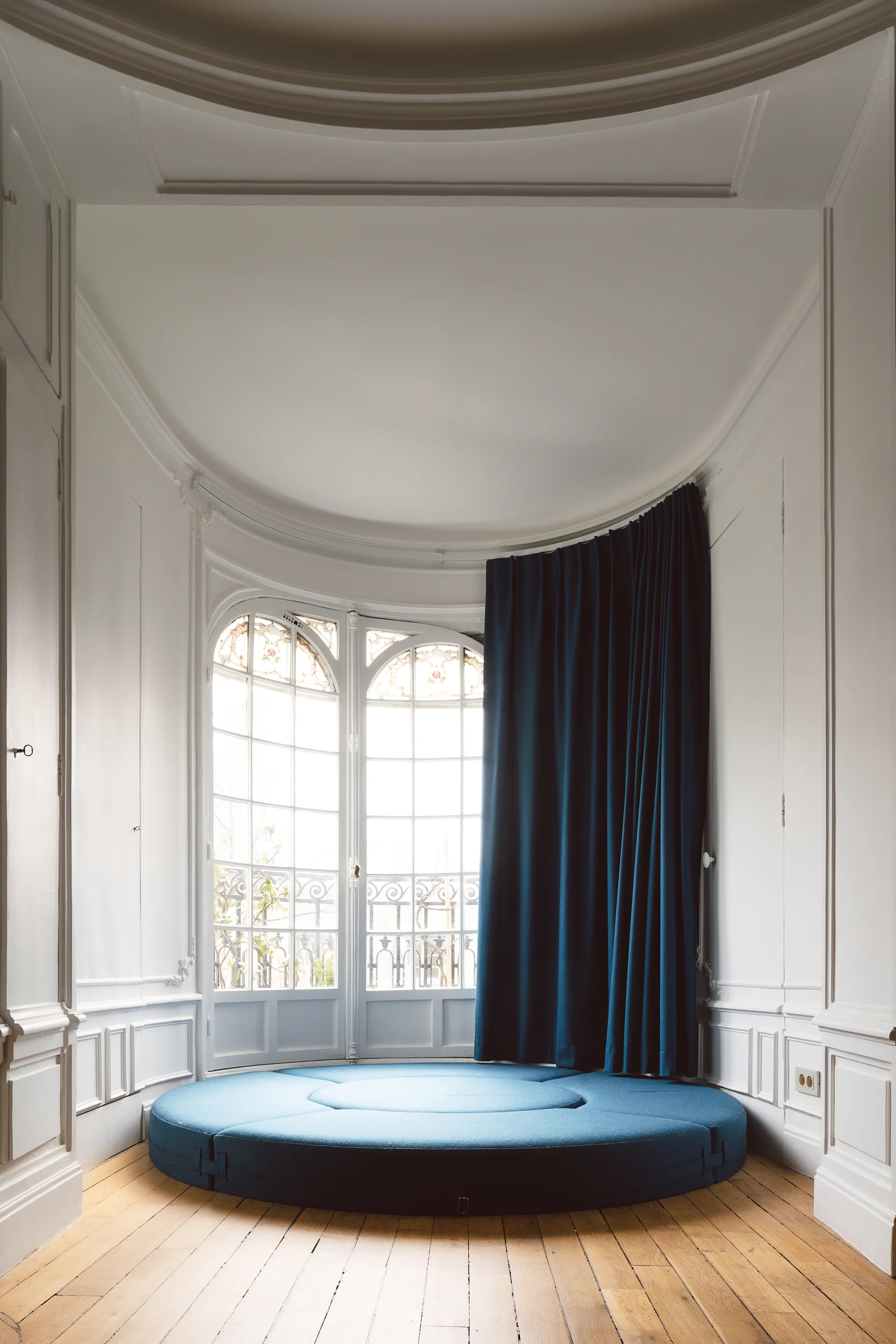 A circular blue upholstered platform rests on wood flooring inside a curved alcove featuring white paneled walls and arched windows.