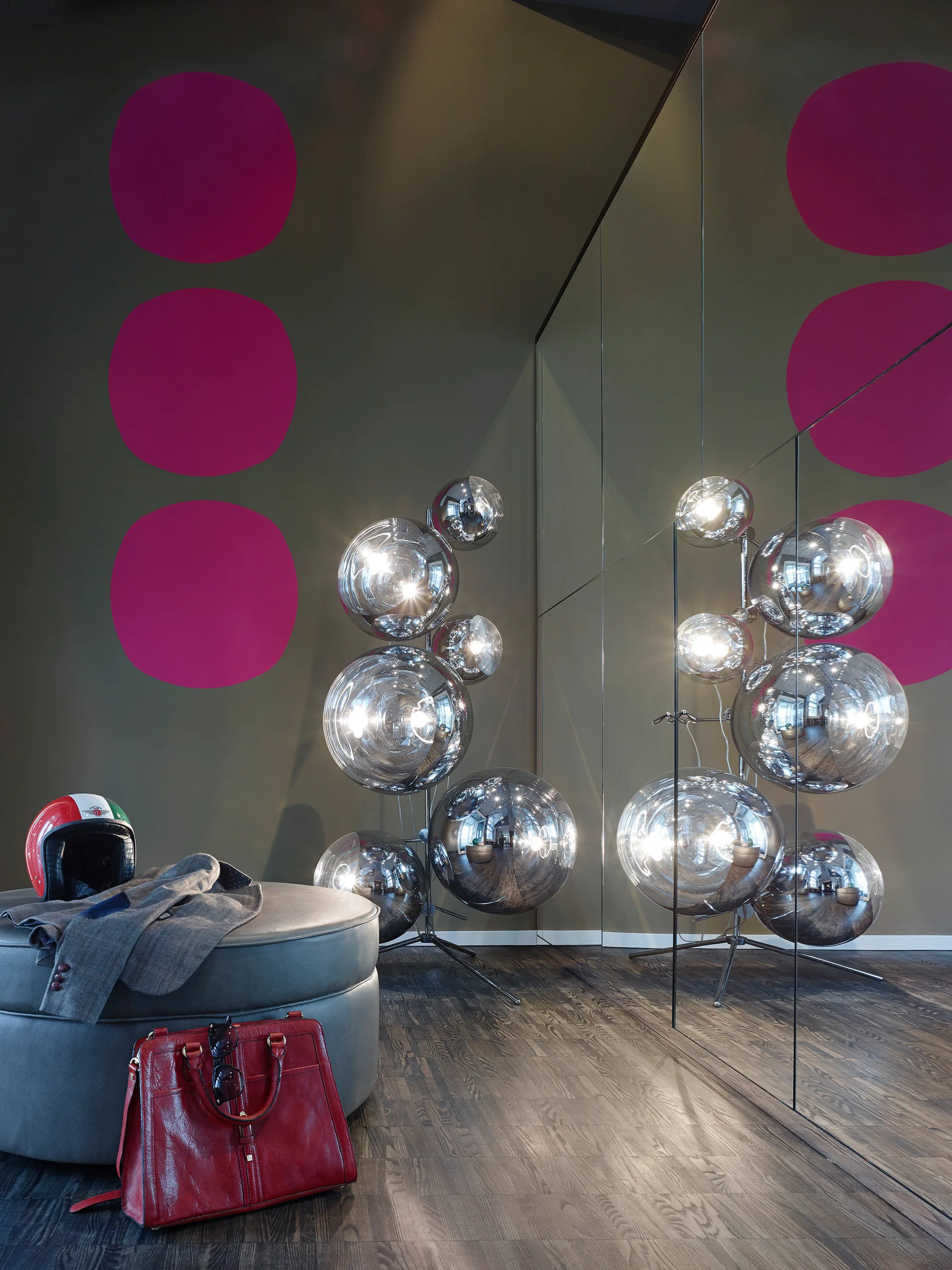 A sculptural chrome sphere floor lamp stands next to a mirrored wall in a room with a round grey leather ottoman and dark wood flooring.
