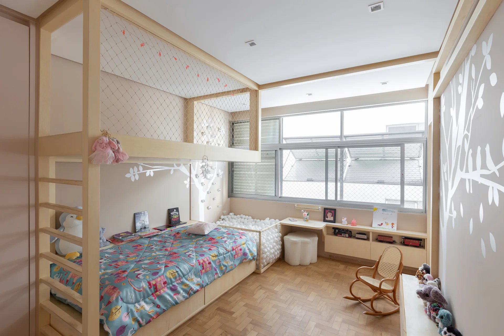 A children's bedroom featuring a custom light wood loft bed, a corner ball pit, a low storage console beneath a wide window, and parquet flooring.