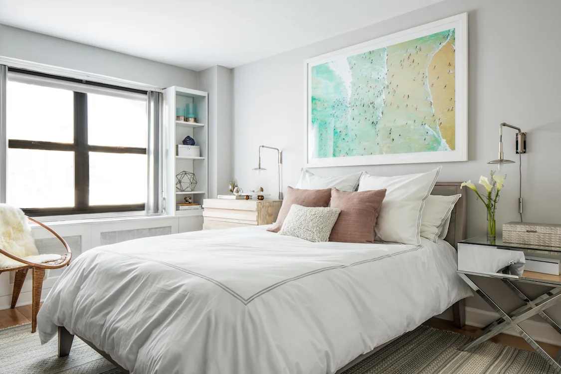 A bright grey bedroom features a bed with white and mauve bedding, a large framed beach photograph on the wall, and a mirrored nightstand.