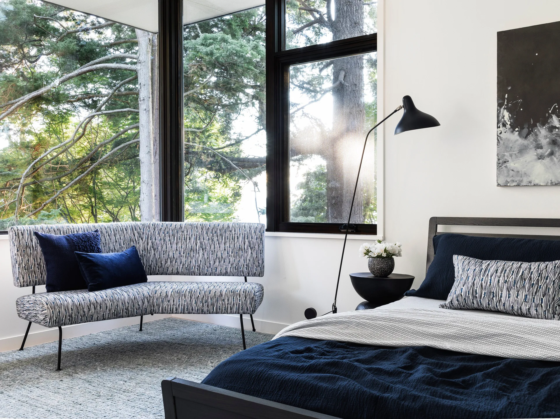 A contemporary bedroom featuring a navy blue bed, a curved patterned sofa, and large black-framed windows overlooking tall green trees.