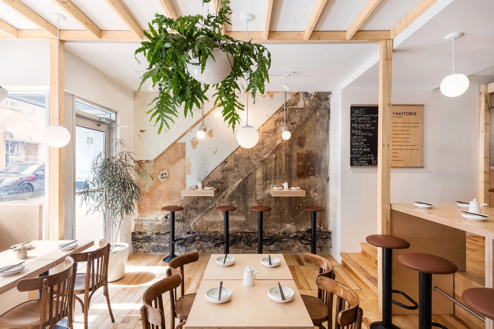 A minimalist restaurant dining area features light wood tables, curved wooden chairs, globe pendant lights, and a diagonal distressed plaster accent wall.