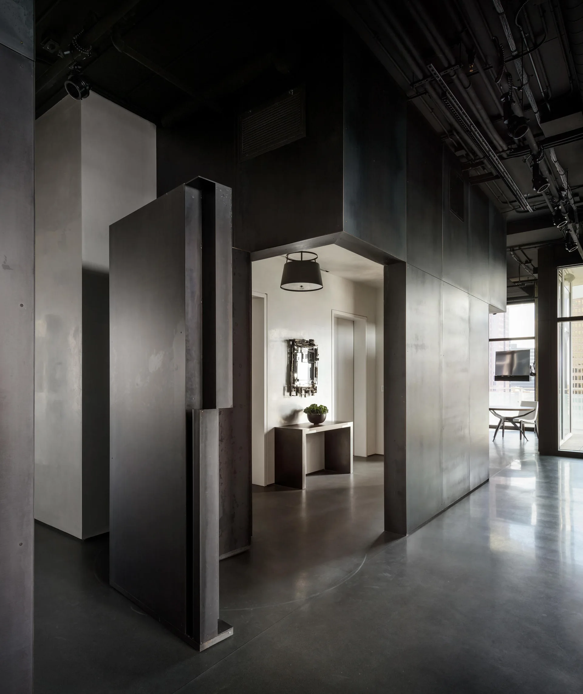 An industrial entryway featuring dark matte steel wall panels, a polished gray floor, an exposed black ceiling, and a white hallway in the background.