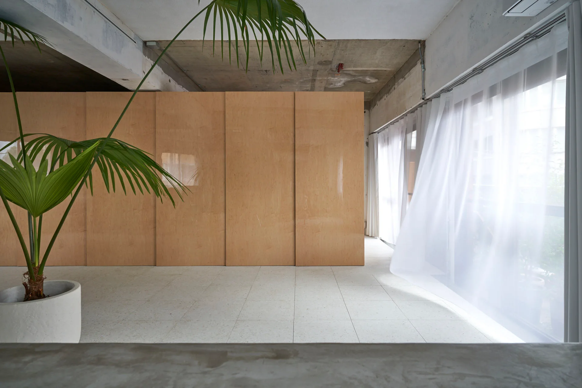 A minimalist interior featuring large light wood plywood partition panels, an exposed concrete ceiling, white tile floors, and sheer curtains.