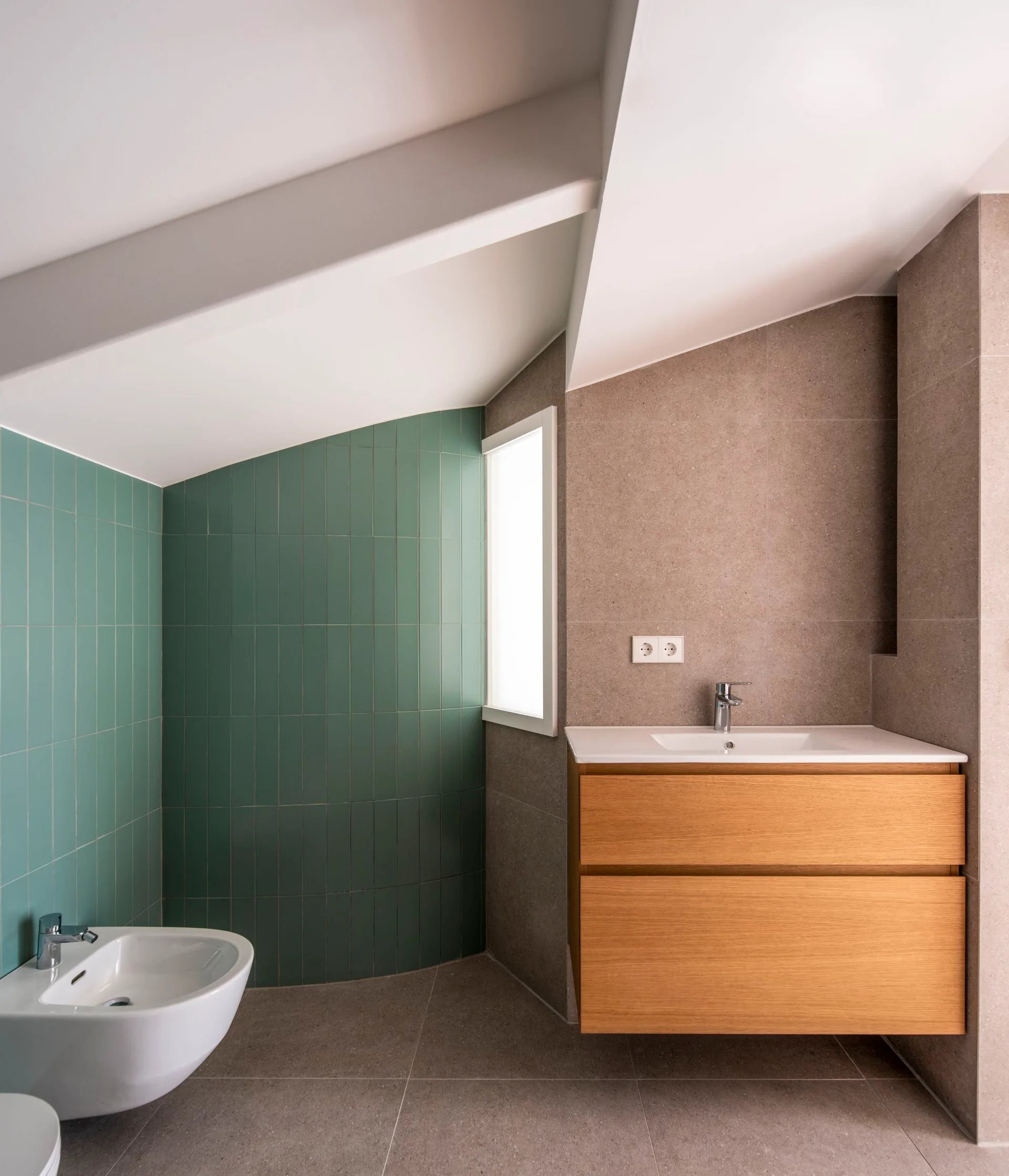 A modern attic bathroom featuring a floating wood vanity with an integrated sink, vertical green wall tiles, and a white bidet under a slanted ceiling.