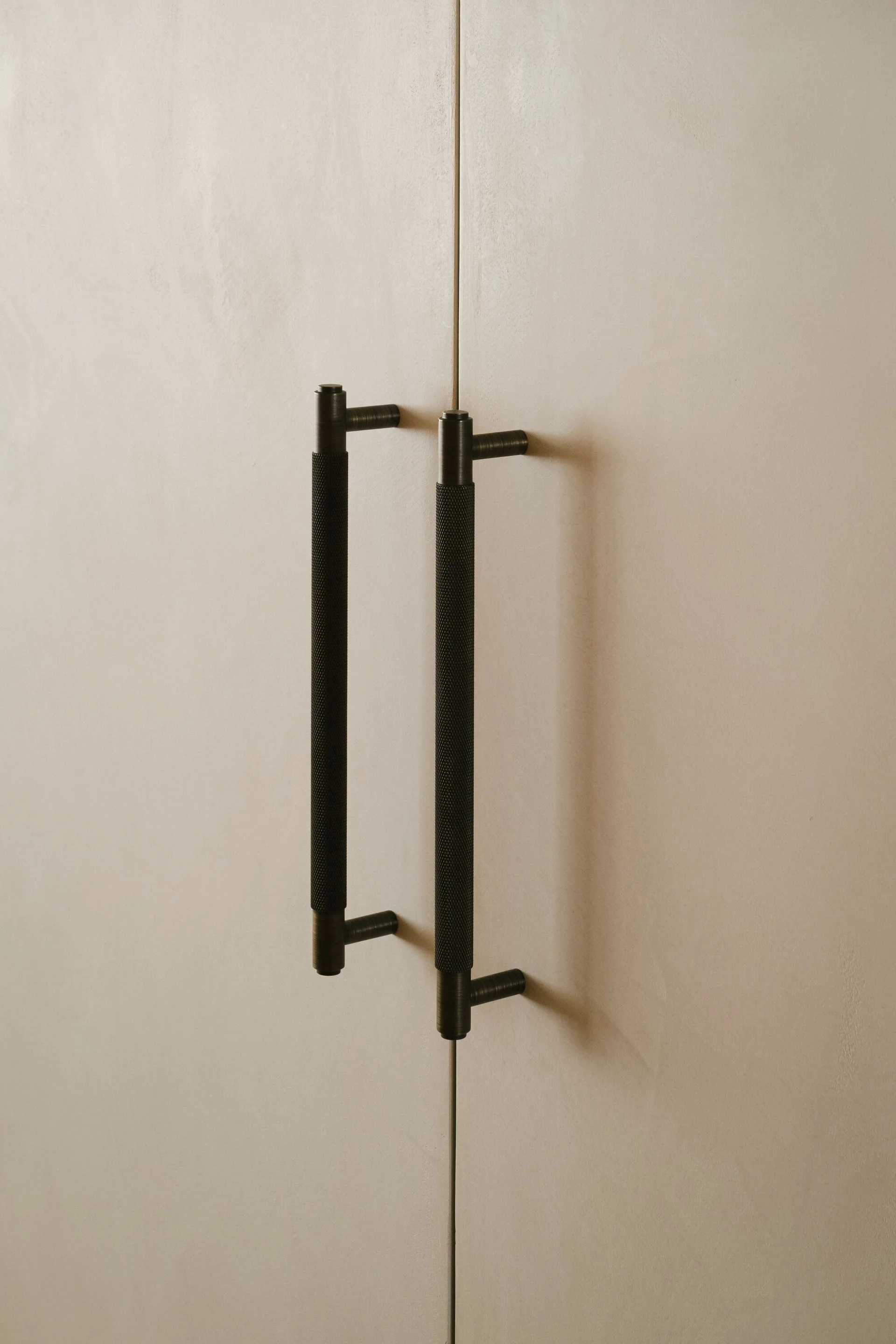 A close-up of textured dark gray knurled metal door handles mounted vertically on minimalist light beige matte cabinet doors.
