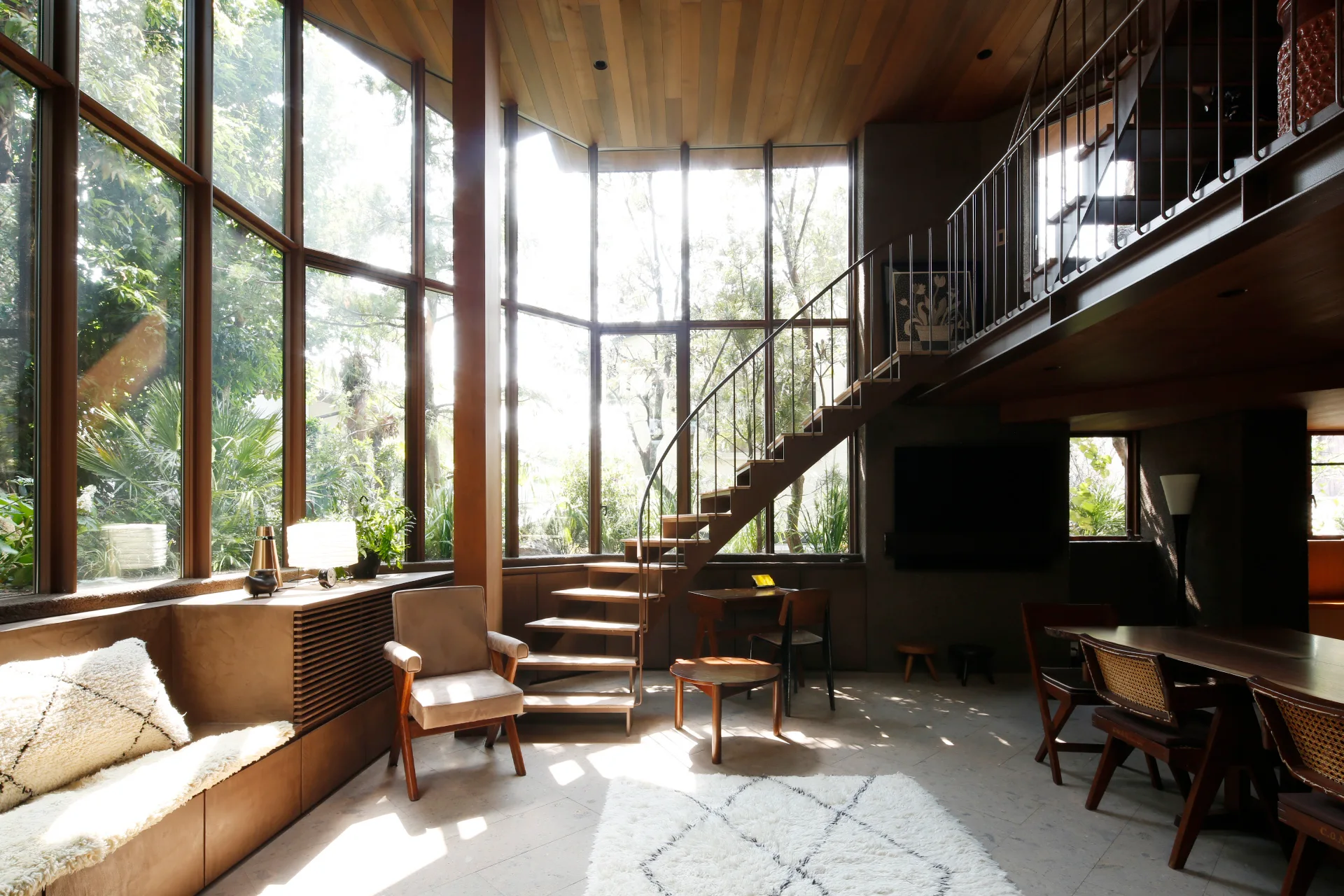 A sunlit mid-century modern living room features a wood-paneled ceiling, a curved staircase, large floor-to-ceiling windows, and low wooden furniture.