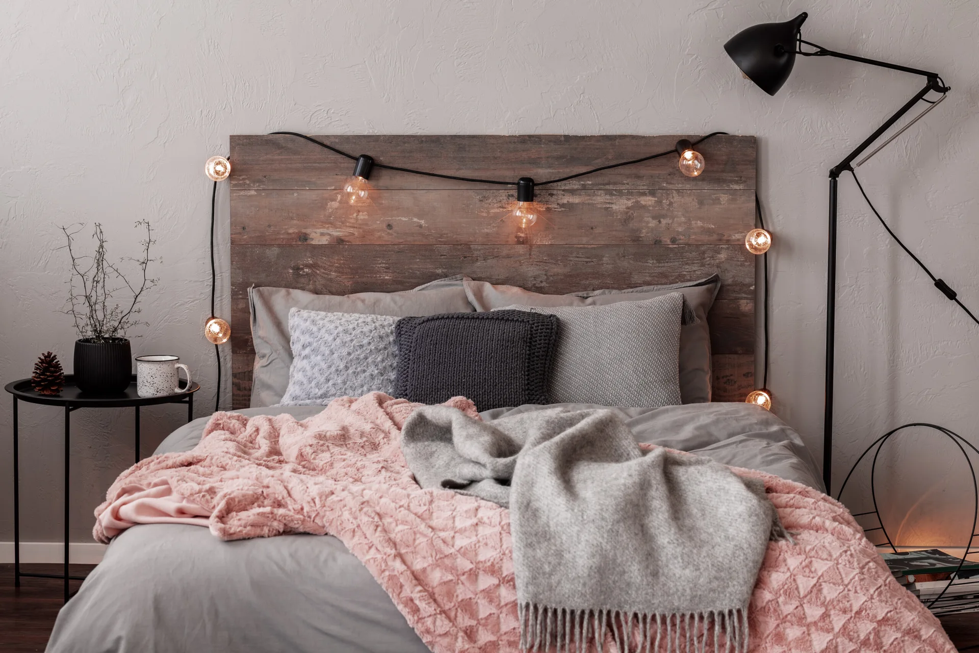 A bed with a rustic wood plank headboard, grey and pink blankets, draped string lights, and a black bedside table against a textured grey wall.