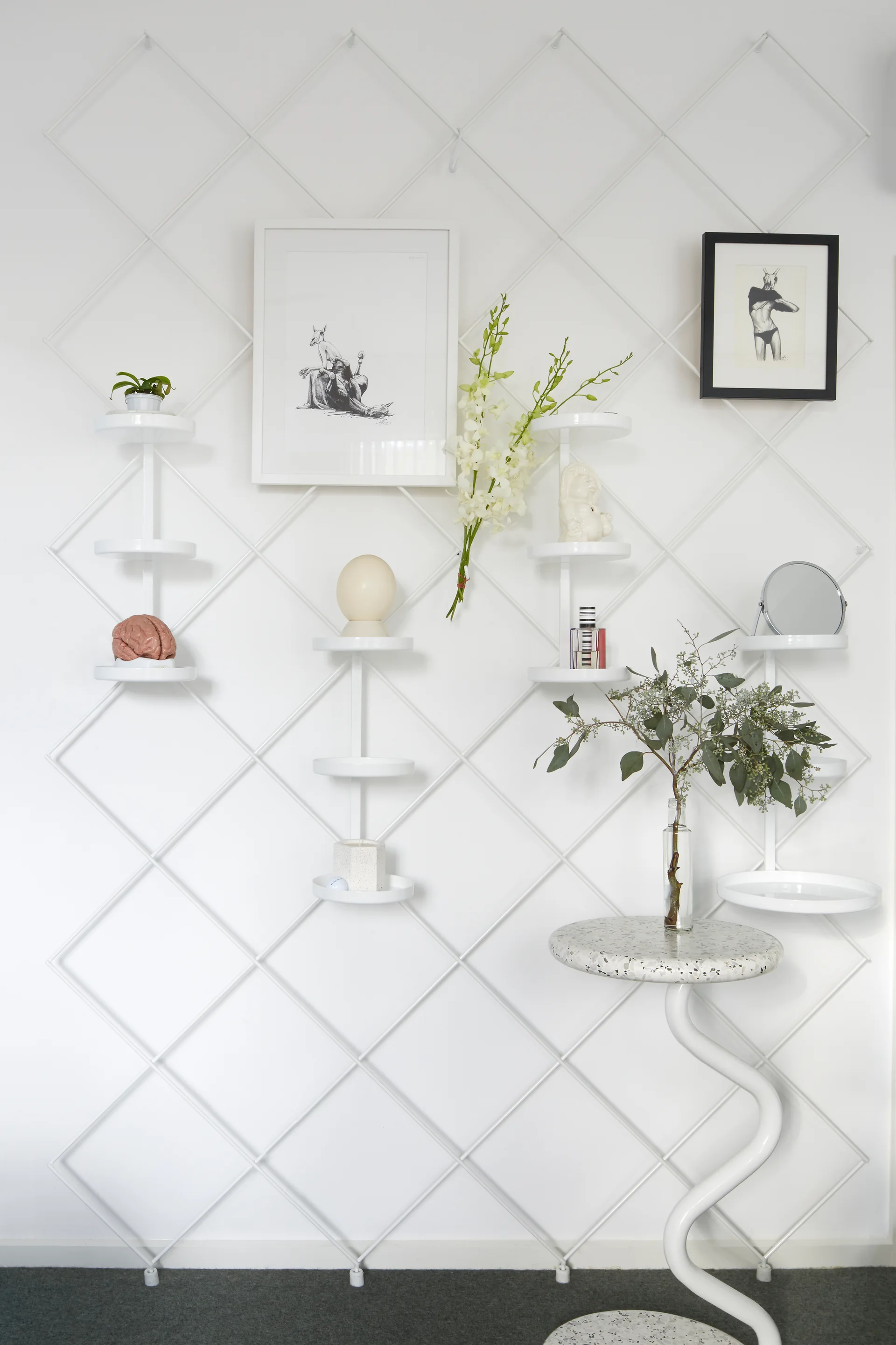 A white metal diamond-grid wall shelving unit displays small decor objects next to a white terrazzo-topped side table with a curved leg.