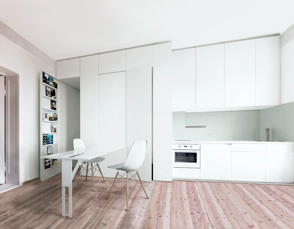 A minimalist white kitchen features handleless cabinets, a light green tiled backsplash, and a slim dining table set on pale wood plank flooring.