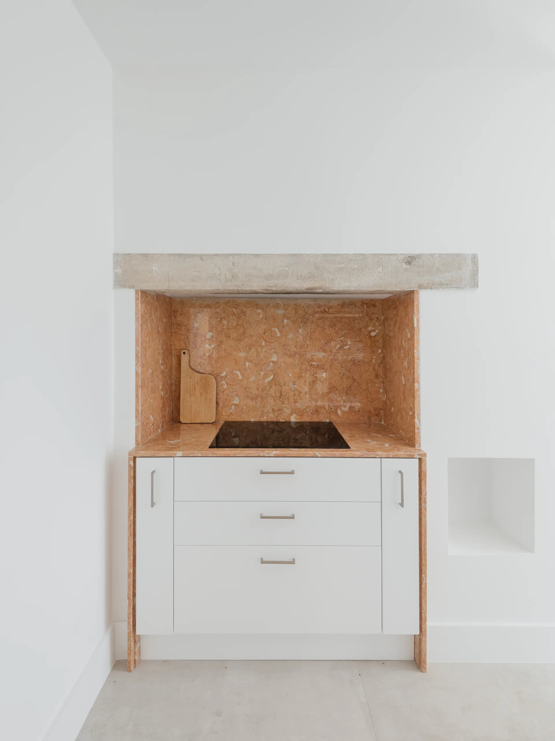 A minimalist kitchen alcove features a white drawer cabinet with a black cooktop, a peach marble backsplash, and a rough stone lintel above.