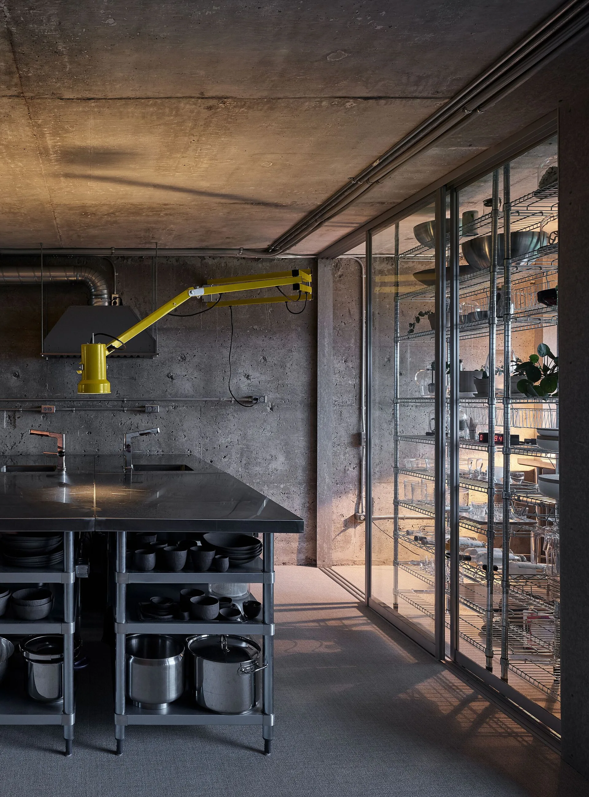 An industrial kitchen features raw concrete walls, a stainless steel island, a yellow adjustable lamp, and a glass-enclosed pantry with wire shelving.