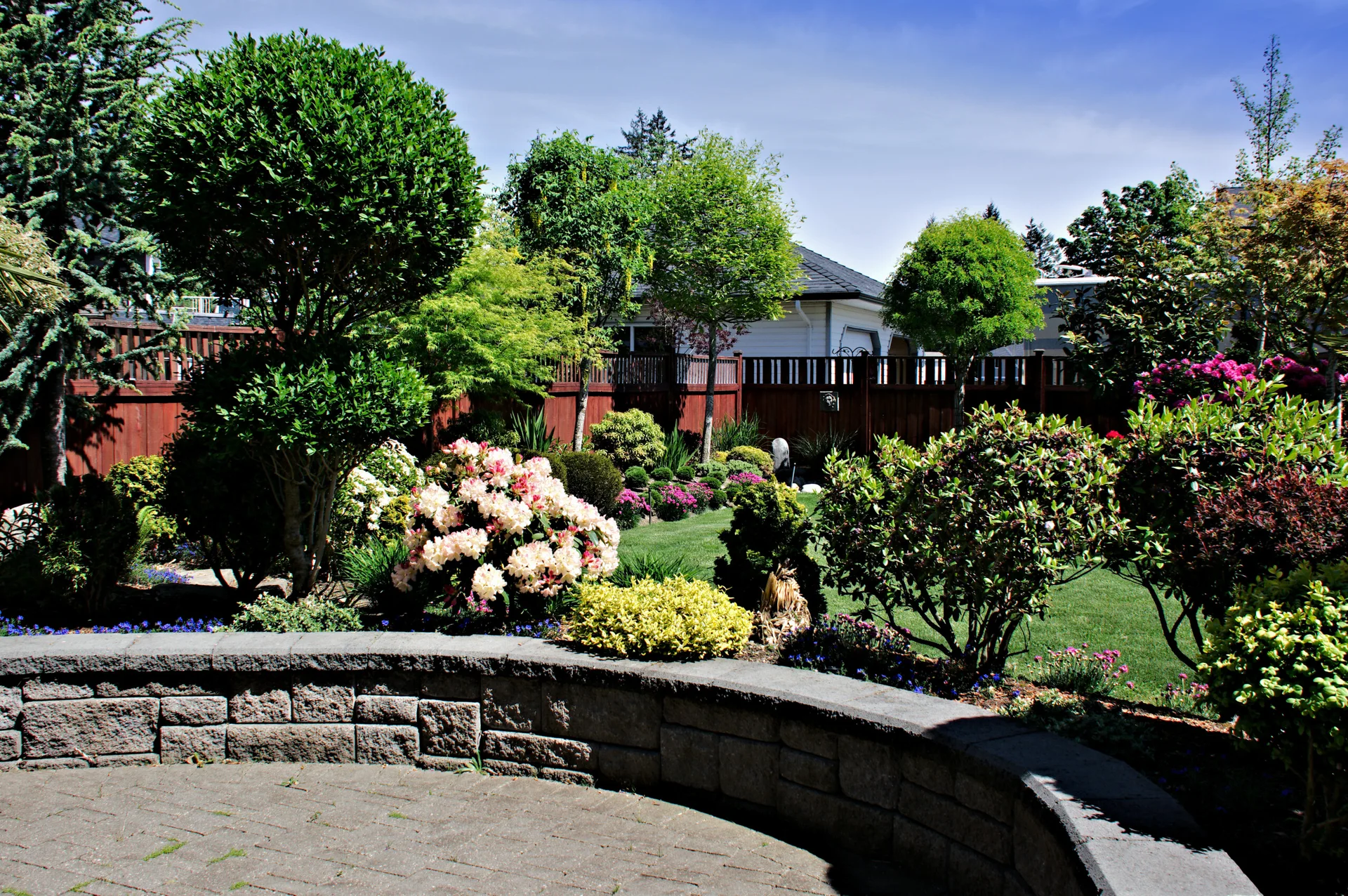 A curved gray stone retaining wall separates a paved patio from a lush green lawn filled with colorful flowering shrubs and a wooden fence.