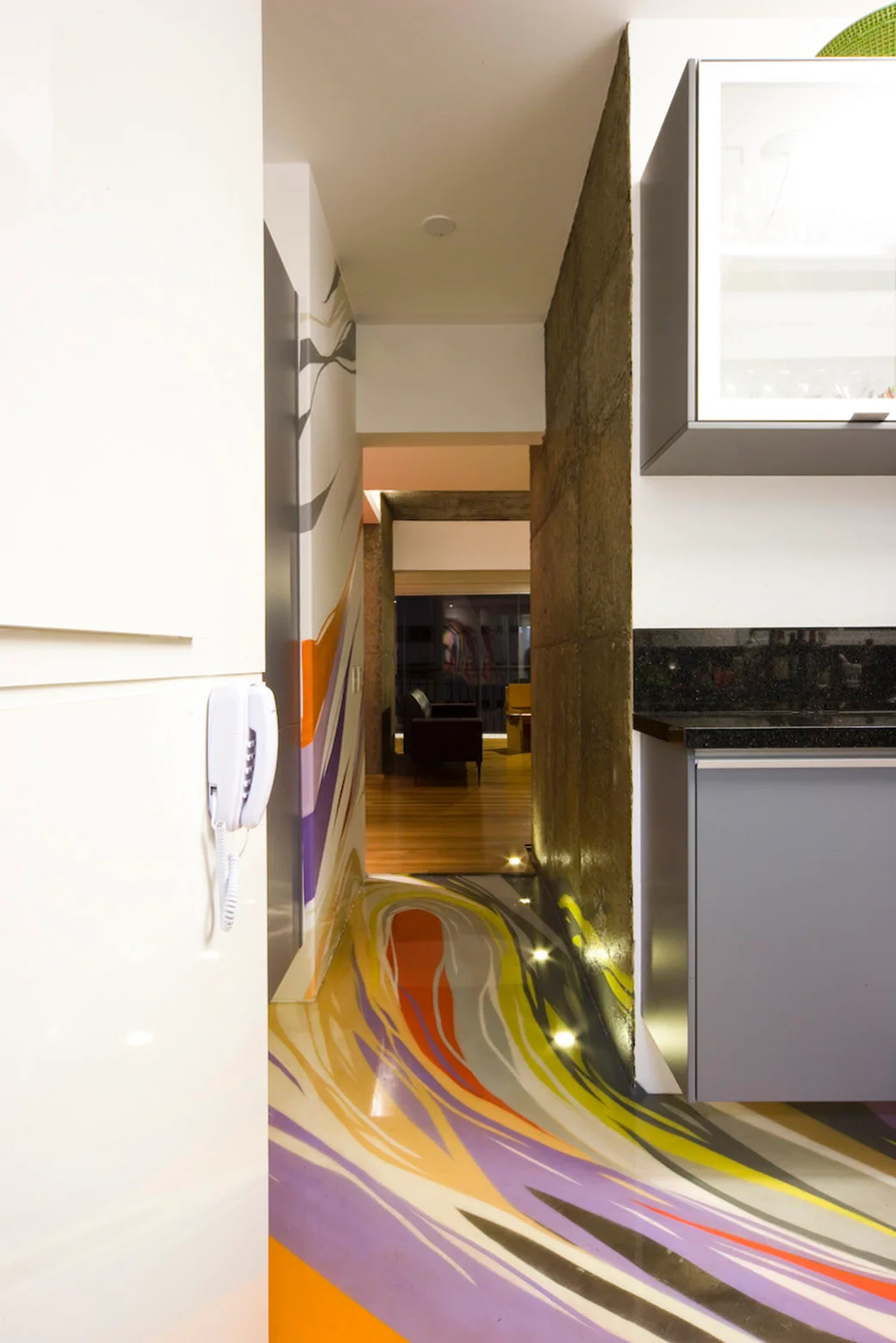 A modern hallway features a glossy, multi-colored swirled floor, a textured grey concrete wall highlighted by floor spotlights, and grey cabinetry.