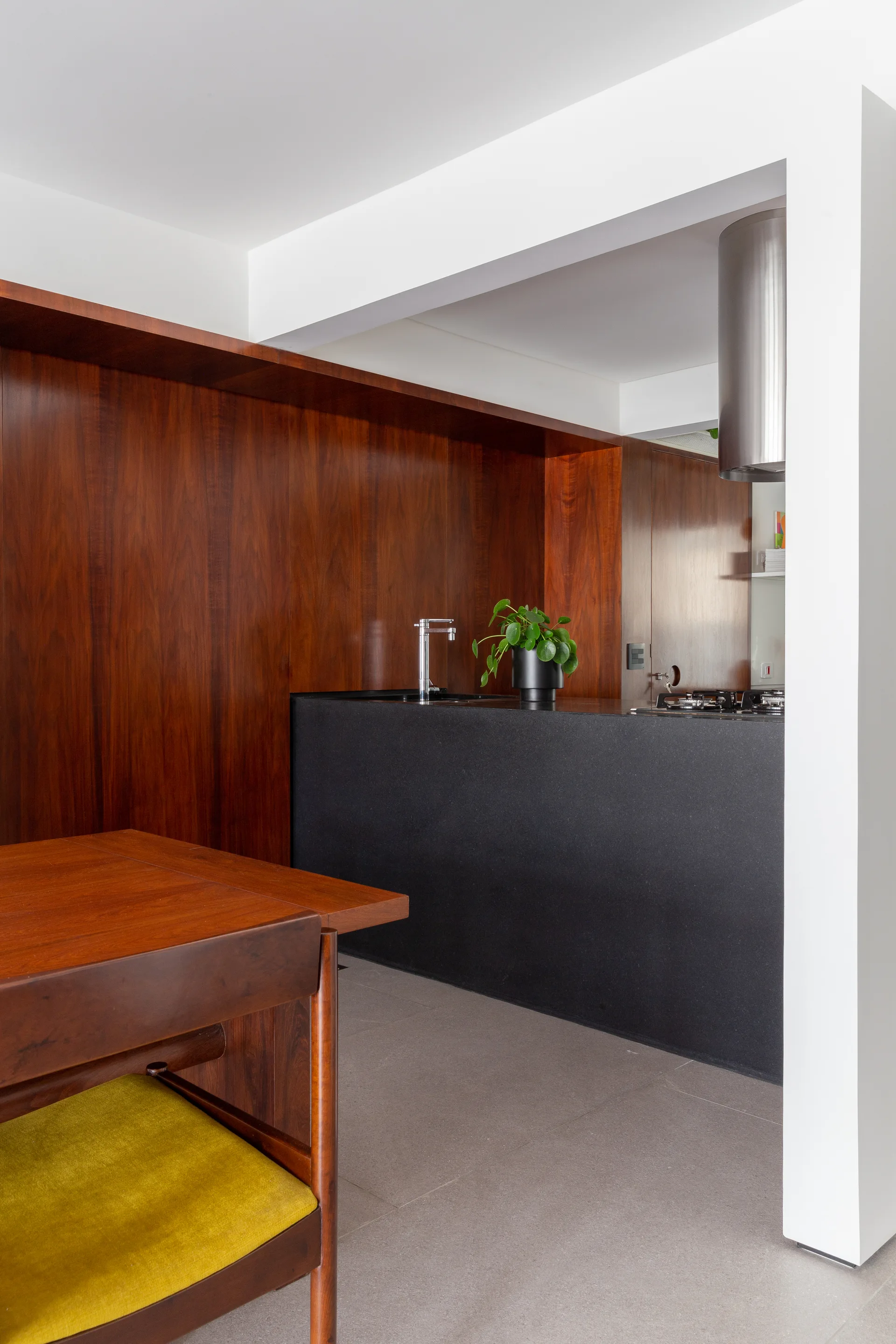 A modern kitchen featuring rich wood wall paneling, a matte black island with a faucet, and a wooden dining chair with a chartreuse seat.