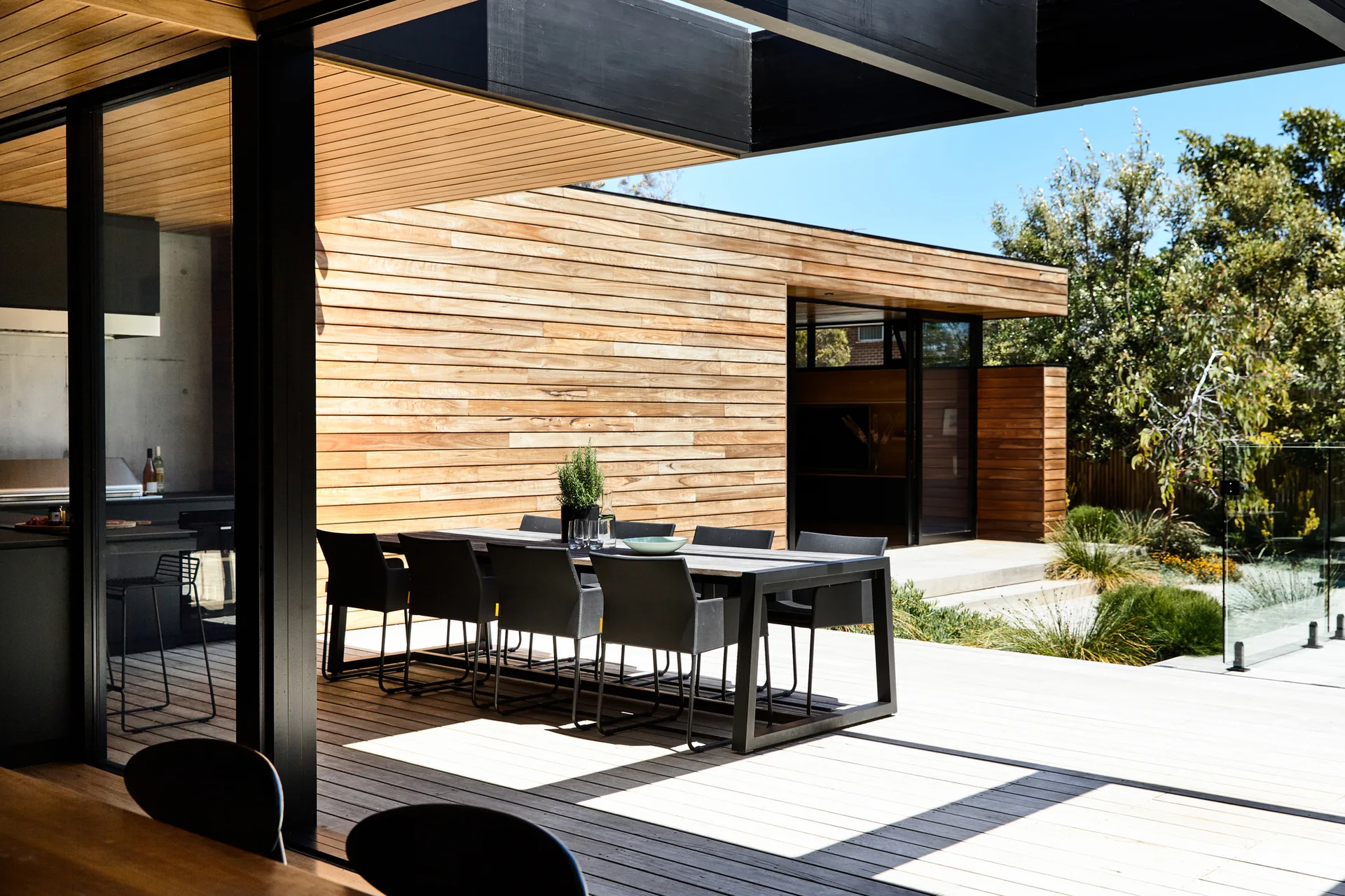 A black dining set sits on a wooden deck next to a house with horizontal timber cladding and large sliding glass doors.