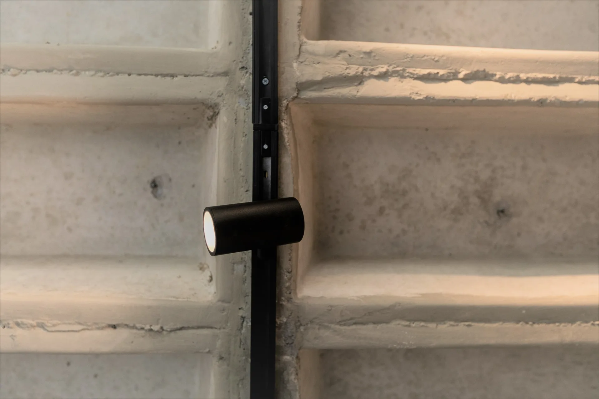 A black track lighting fixture with a single illuminated spotlight mounted vertically on a rough gray concrete grid wall.